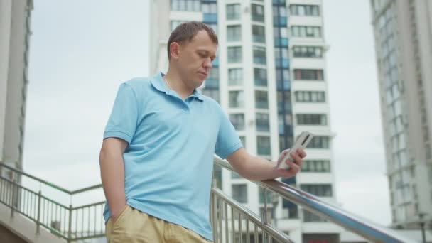 Homem com um telefone celular em suas mãos, fica nas escadas, prédios de fundo — Vídeo de Stock