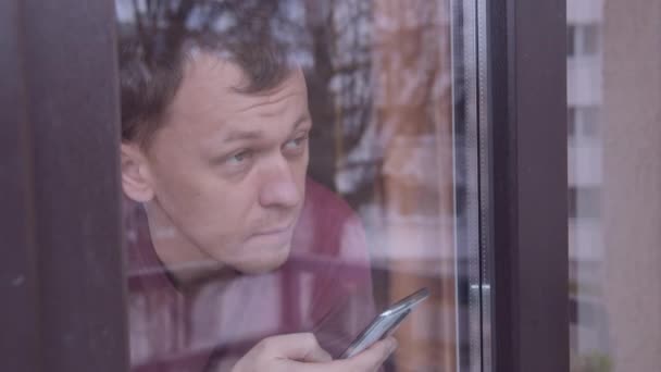 Чоловік за вікном, дивлячись на вулицю і використовуючи мобільний телефон, стеження за камерою — стокове відео