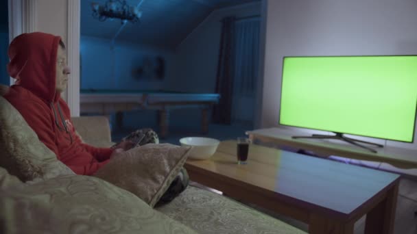 Een man zit 's avonds op een bank met een mobiele telefoon voor een groene tv — Stockvideo