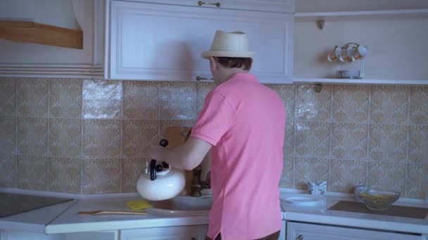 Hombre con sombrero está bailando en la cocina vertiendo agua en un hervidor de agua, buen humor — Vídeo de stock