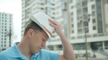 Adam sokakta dikilirken şapka takıyor, binalar arka planda.