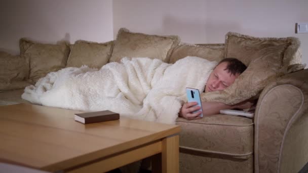 L'homme est allongé sur le canapé la nuit à regarder la télévision, tient un téléphone dans ses mains, l'éclairage du soir — Video