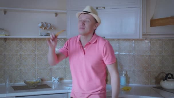 Een man met een roze T-shirt en een hoed danst en zingt in de keuken, goed humeur — Stockvideo