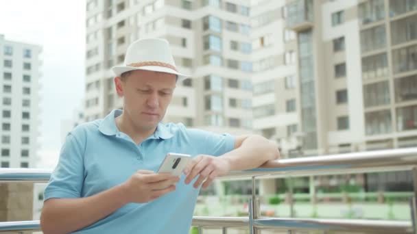 Гражданин в шляпе просматривает свой мобильный телефон, пишет текстовое сообщение, стенды, фон городских зданий — стоковое видео