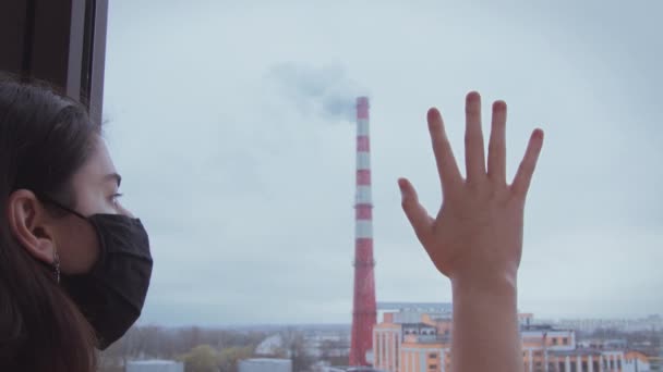 Dívka v černé masce se podívá z okna a položí ruku na sklo. Průmyslové potrubí za oknem — Stock video