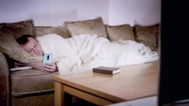 Un homme est allongé sur le canapé la nuit à regarder la télévision, tient un téléphone dans ses mains, l'éclairage du soir — Video