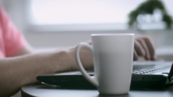 Manos masculinas escribiendo en un teclado portátil, escribiendo texto, junto a una taza con café, fondo borroso — Vídeos de Stock