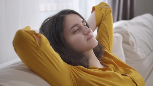 Chica feliz relajándose en el sofá con los ojos cerrados, las manos detrás de la cabeza, sentado en casa. Soñador — Vídeos de Stock