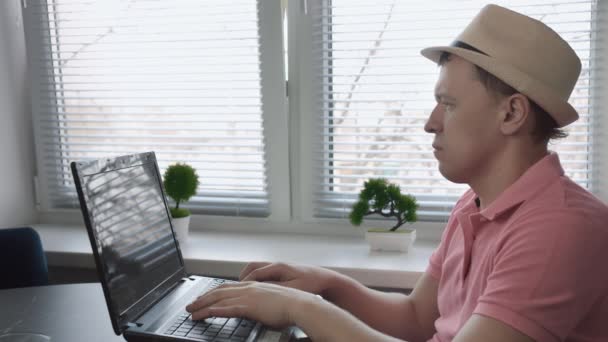 Hombre freelancer en sombrero sentado en la mesa y escribir texto en el ordenador portátil, fondo de ventana — Vídeos de Stock