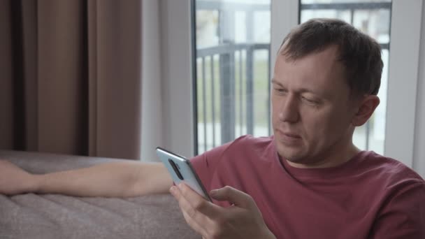 Homem triste ler mensagens de texto navegar na Internet em Smartphone Sentado no sofá — Vídeo de Stock