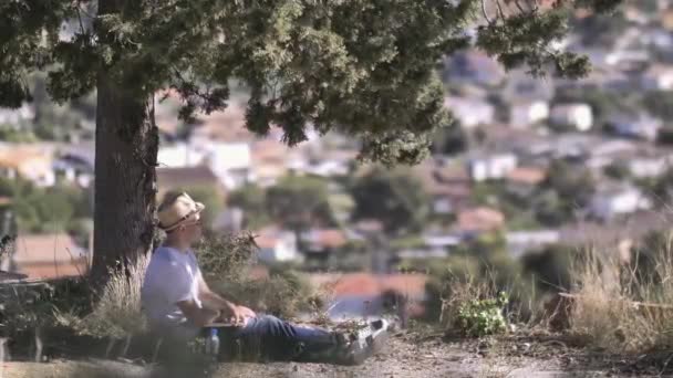 Молодой человек в шляпе и с ноутбуком сидит под деревом — стоковое видео