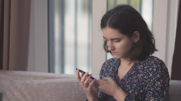 Portrait d'une fille avec un téléphone portable dans les mains, assise sur le canapé, fond de fenêtre — Video