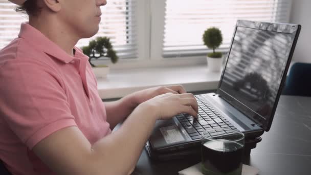 Freelancer masculino sentado en la mesa y escribiendo texto en el portátil, fondo de ventana, oficina en casa, seguimiento de la cámara — Vídeos de Stock