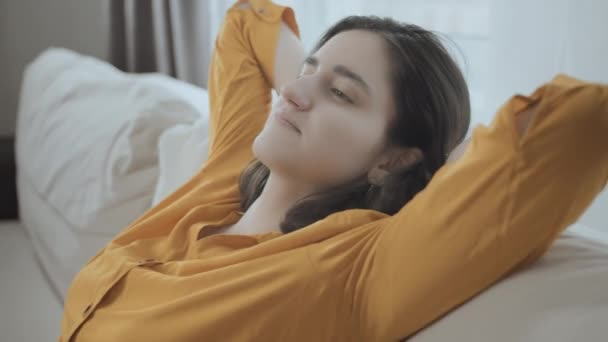 Chica en el sofá, sentada cómodamente con las manos detrás de la cabeza, con una camisa amarilla, fondo de ventana. Disfrutando del momento — Vídeos de Stock