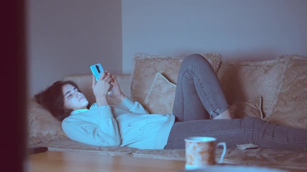 Adolescente chica se encuentra en la noche cerca de la televisión en el sofá con un teléfono móvil — Vídeo de stock