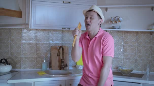 Een man met een roze T-shirt en een hoed danst en zingt in de keuken — Stockvideo
