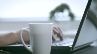 Erkek eller dizüstü bilgisayarda yazı yazıyor, yazı yazıyor, kahve bardağının yanında, arkaplan bulanık.