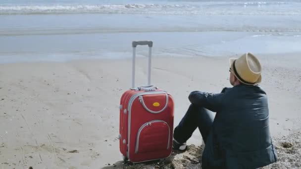 Tourist mit Hut und rotem Koffer sitzt am Strand, Urlaubskonzept — Stockvideo