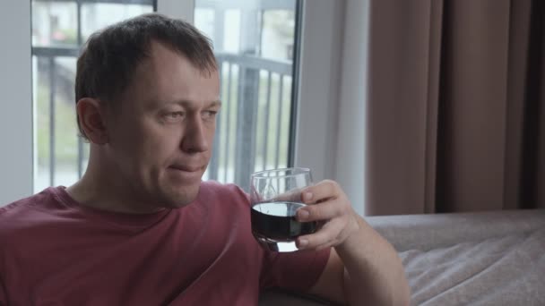 Одинокий человек, пьющий из стакана, сидящего на диване, на фоне окна — стоковое видео