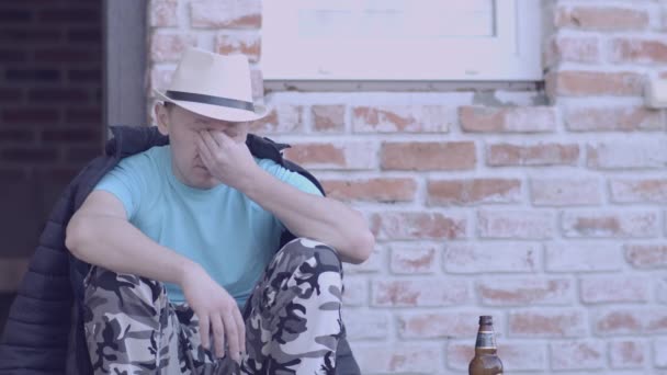 Młody człowiek w kapeluszu siedzi na ganku domu, obok butelki piwa. — Wideo stockowe