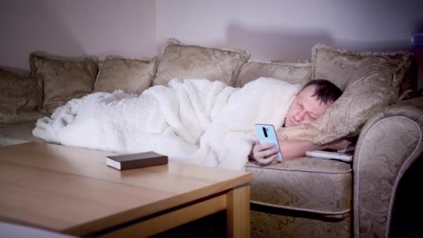 Un homme seul, allongé sur le canapé la nuit, regarde la télé, tient un téléphone dans ses mains — Video