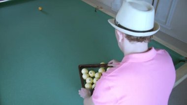 Bilardo oynayan, pembe tişörtlü ve şapkalı bir adam üçgende top topluyor.