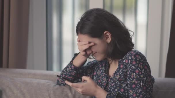 Brünettes Mädchen lacht, liest Nachrichten in ihrem Handy, sitzt auf dem Sofa, Hintergrund des Fensters — Stockvideo