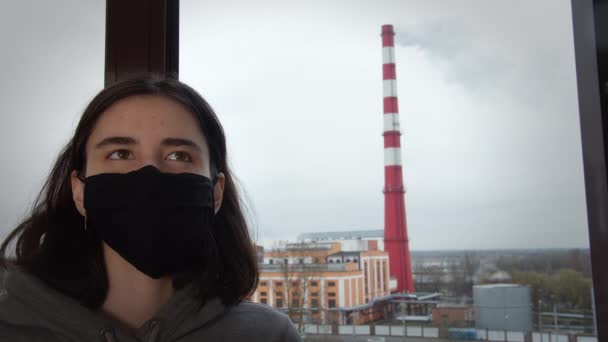 Дівчина в захисній масці на тлі диму з труби через скло, стеження за камерою — стокове відео