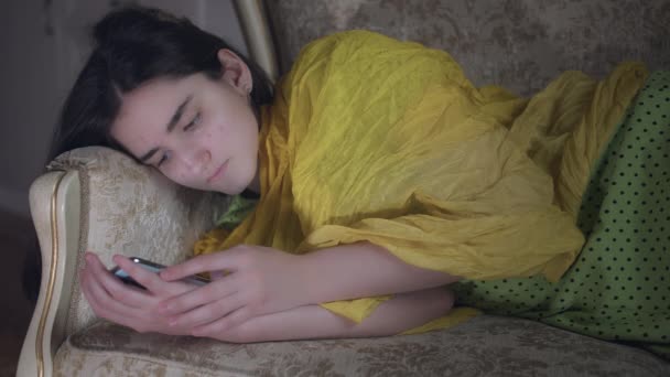 A brunette girl lies in the evening on the couch with a mobile phone — Stock Video