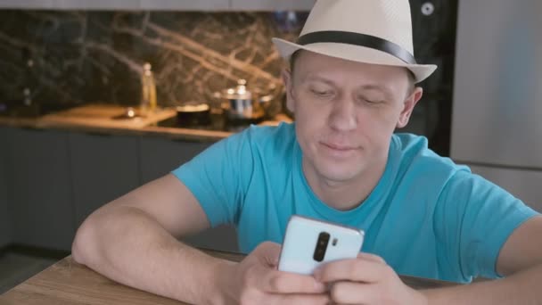 Πορτρέτο ενός ελκυστικού άνδρα που σε ένα λευκό καπέλο κάθεται σε ένα τραπέζι χρησιμοποιεί ένα κινητό τηλέφωνο — Αρχείο Βίντεο