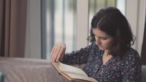 Brünettes Mädchen liest ein Buch, während es auf dem Sofa sitzt, Fensterhintergrund — Stockvideo