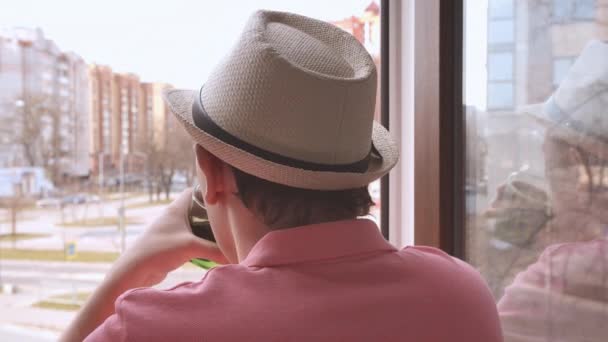 Молодий чоловік у капелюсі, що стоїть у віконному отворі, дивиться на вулицю і п'є напій зі склянки — стокове відео