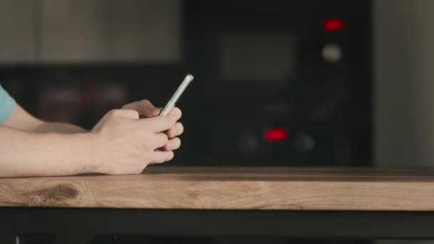 Seorang pria dengan ponsel di tangannya berdiri di dekat meja kayu, menggunakan aplikasi seluler, latar belakang dapur — Stok Video