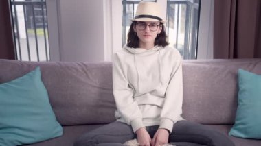 Gözlüklü ve şapkalı bir kız, bacak bacak üstüne atmış bir şekilde koltukta oturuyor, doğrudan kameraya bakıyor, arka planda pencere var.