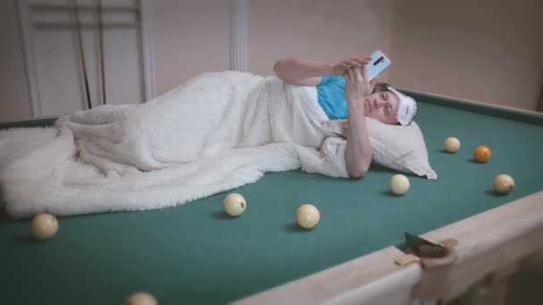 Un hombre con una máscara de dormir se encuentra en una mesa de billar entre las bolas cubiertas con una manta, utiliza un teléfono móvil — Vídeo de stock