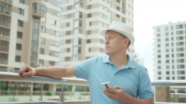 Şapkalı ve elinde telefon olan bir adam, şehir caddesinde metal bir çitin yanında duruyor, kamera takip ediyor.