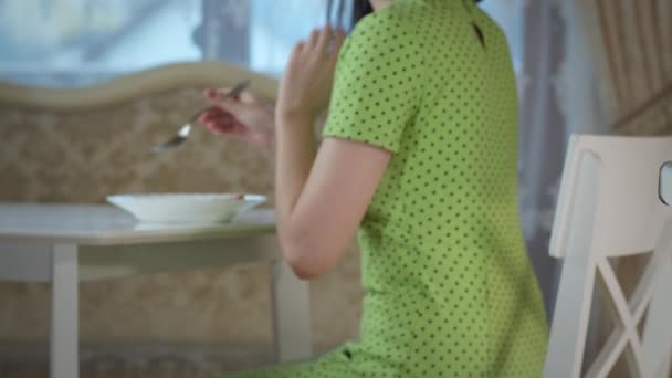 Dziewczyna w zielonej sukience je śniadanie siedząc przy białym stole w domu — Wideo stockowe