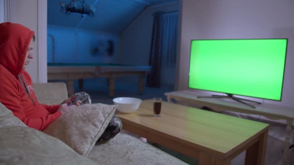 Νεαρός άνδρας βλέποντας τηλεόραση και χρησιμοποιώντας το κινητό τηλέφωνο, κάθεται στον καναπέ στο σπίτι το βράδυ, chroma κλειδί — Αρχείο Βίντεο