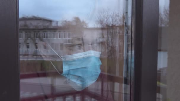 Человек в маске у окна, смотрит на улицу через стекло, отражение улицы на стекле, слежение за камерой — стоковое видео