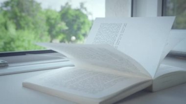 Açık bir pencerenin pencere pervazında bir kitap uzanıyor, güneş ışınları sayfalarda, yakın plan.