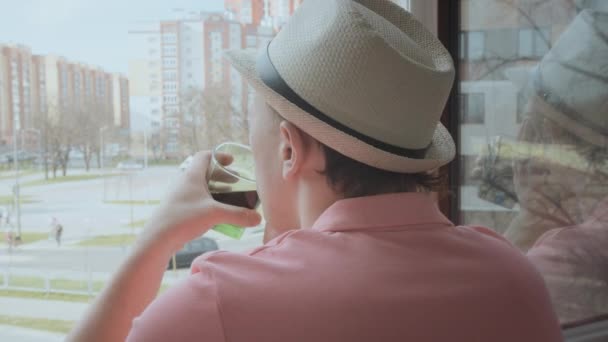 Homem com um copo na mão fica em uma janela aberta, bebe uma bebida e olha para a rua, vista para trás — Vídeo de Stock
