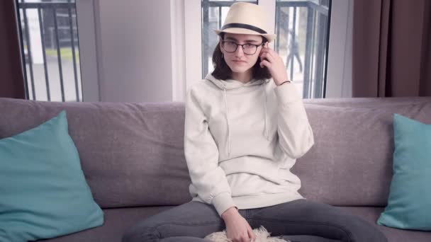 Mädchen mit Brille und Hut blickt direkt in die Kamera, die im Schneidersitz auf der Couch sitzt — Stockvideo