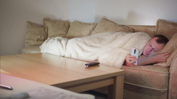 Ein Mann, der nachts auf der Couch am Fernseher liegt und sein Handy benutzt, kopiert Raum — Stockvideo