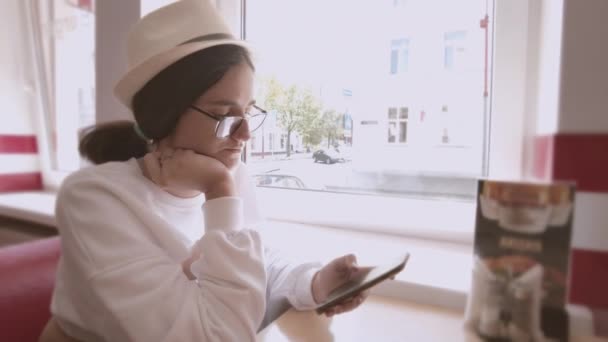 Девушка в шляпе и солнцезащитных очках сидит в кафе, пользуется телефоном, ждет приказа, отслеживает камеру — стоковое видео