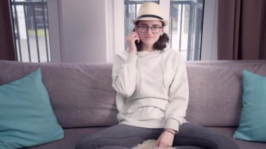 Gözlüklü ve şapkalı güzel kız kanepede bağdaş kurup kameraya bakıyor.