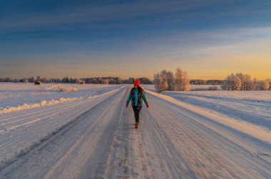 A beautiful solo explorer woman wearing colourful cloth walking on the empty road with snow covered scenic landscape view and sun light from horizon