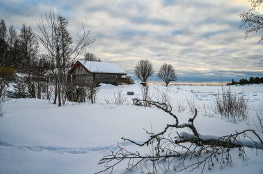 An evening landscape of cloud covered sky and snow covered lake in northern Europe with branches of dead tree and lonely cottage