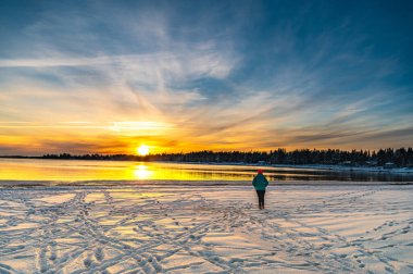 Tek başına gezen kadın kış boyunca karla kaplı bir sahilde günbatımının panoramik manzarasının tadını çıkarıyor.