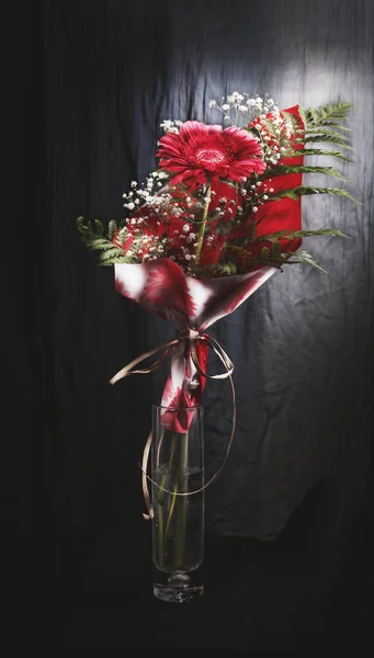A glass vase with a red flower surrounded by wrapping paper on a dark background