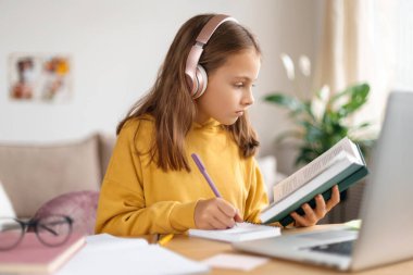 Liseli kız çevrimiçi çalışıyor. Evde eğitim. Uzaktan eğitim. Küçük kız kitap okuyor, ödev yapıyor, uzaktan eğitim için dizüstü bilgisayar ve kulaklık kullanıyor, okul öğretmeniyle online ders alıyor.